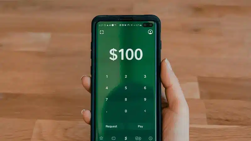 Smartphone affichant l'interface de paiement B-Sharpe avec montant de 100 dollars, clavier numérique et boutons Request/Pay